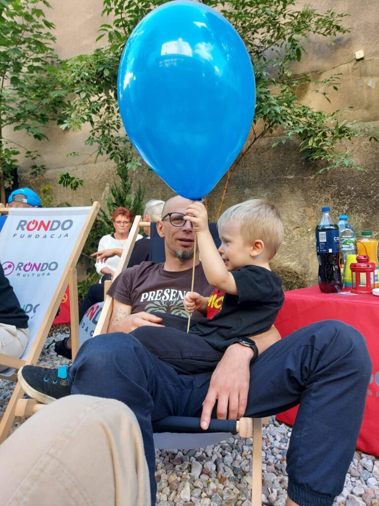 Paweł z Małym Malte na kolanach, który trzyma z radością niebieski balon podczas otwarcia kawiarenki społecznej